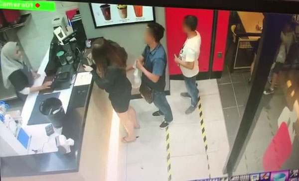 一名女子在柜台前向工作人员点餐，而排在后面的男子便将手伸入女子的手提包。 