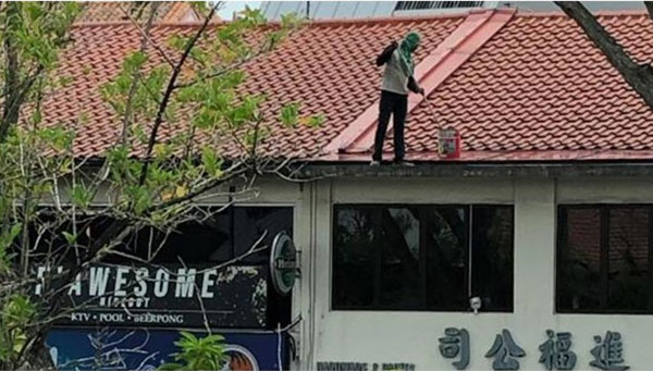  男子一手握刷子，一手提油漆桶，在两层楼店屋的屋顶上来回走动粉刷，对面顾客看得胆战心惊。（受访者提供）