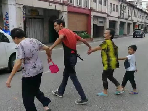 诺西达沙华左右手皆牵着一名婆婆，协助带领她们越过马路。