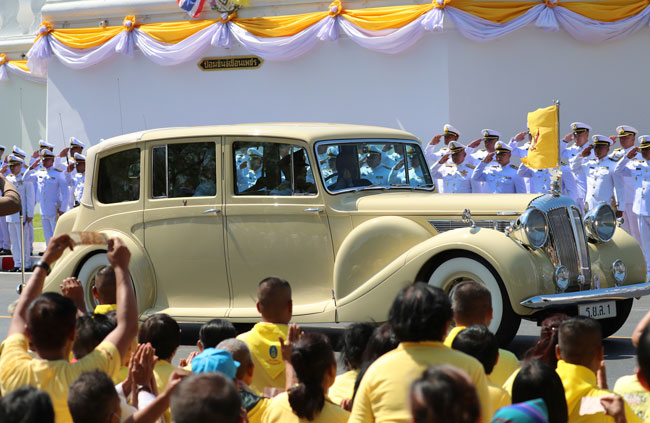 泰王和王后乘坐王家汽车，沿途有官员敬礼，穿上黄衣的民众在旁观看。（美联社）