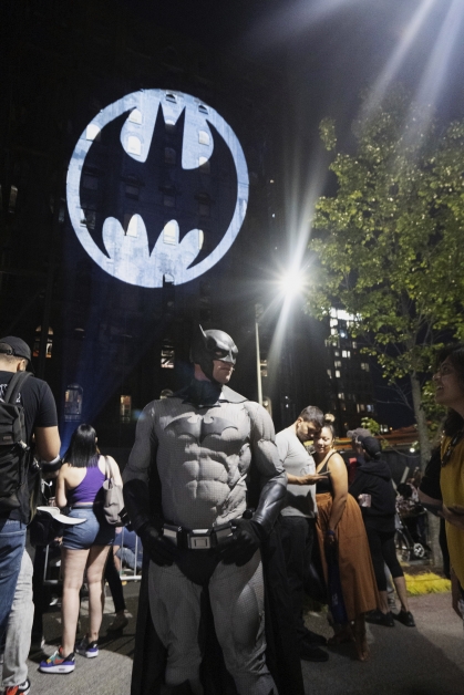 穿上蝙蝠侠图案披风的蝙蝠侠迷在亮起蝙蝠灯的摩天大楼下照相。