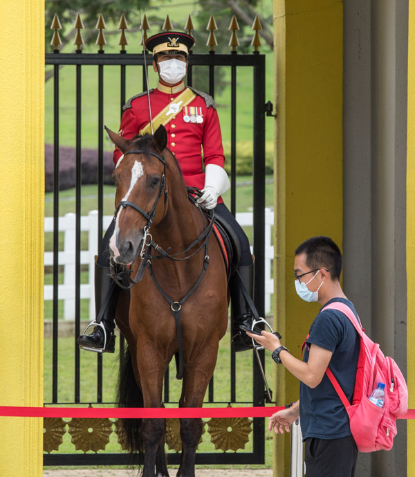 周一，吉隆坡国家王宫外的卫兵戴着口罩骑马，一名旅客在他面前经过。欧新社