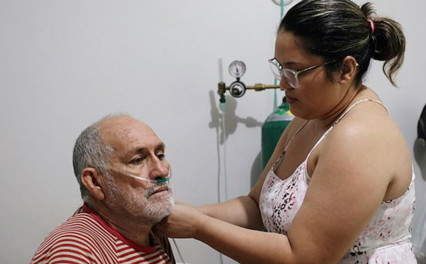 68岁的冠病患者玛格海斯被迫在家中自疗，女儿正为他接驳氧气供应。