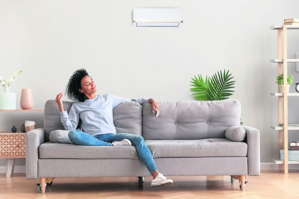 African american relaxed woman sitting on comfortable couch in living room at modern home holds air conditioner remote control enjoying breathing fresh cool air at summer or warm air at winter season